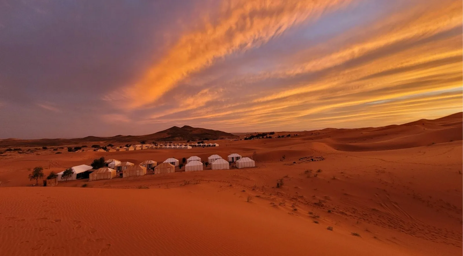 White desert camp tents among orange dunes under a burning sunset sky